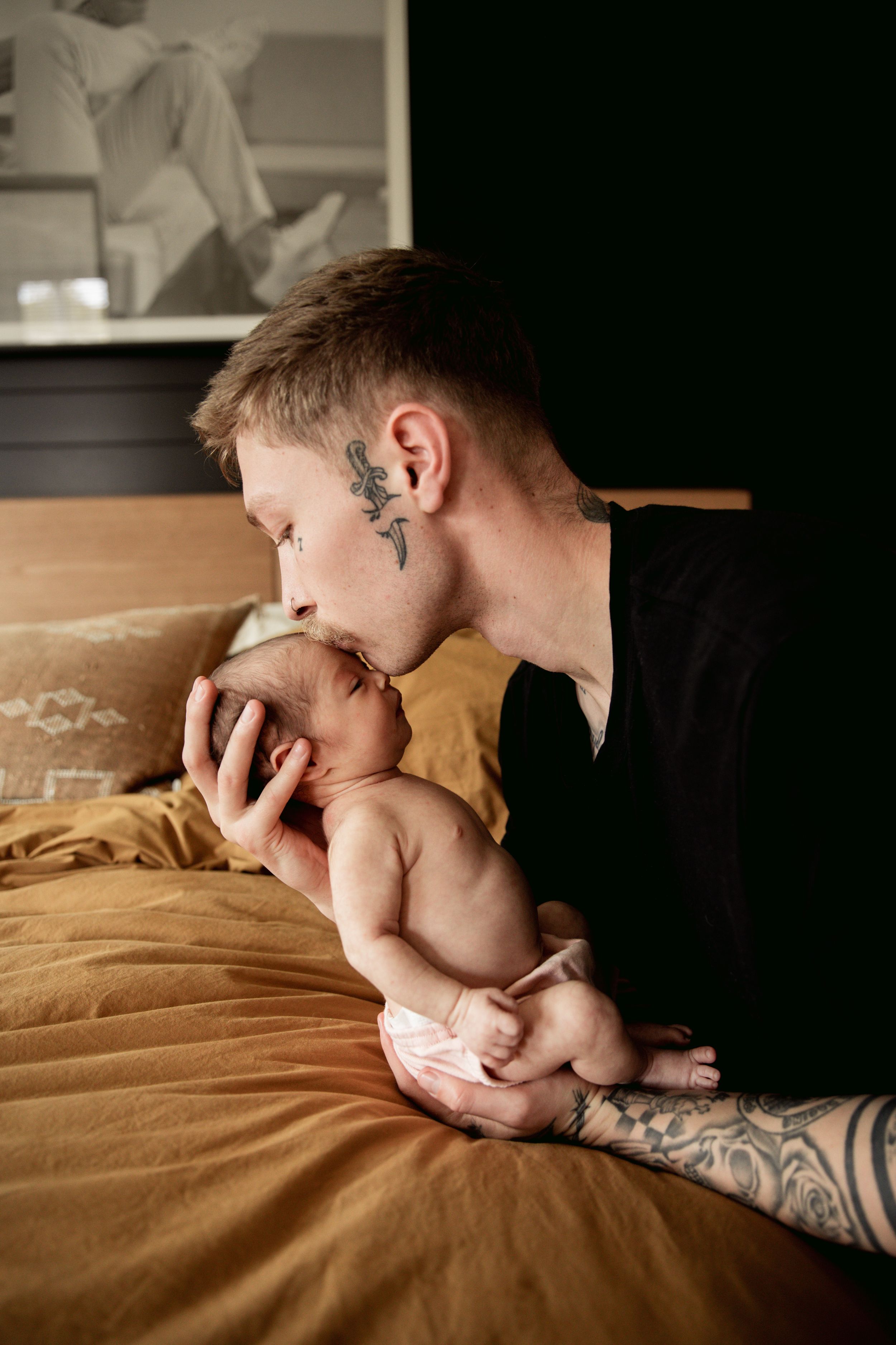 A person with tattoos holding and kissing a baby on an amber-colored bed.