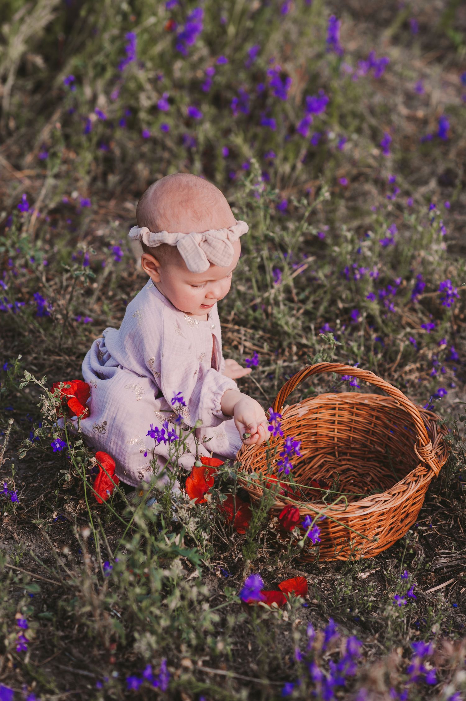 A small child in a light dress sits among purple wildflowers next to a woven basket.