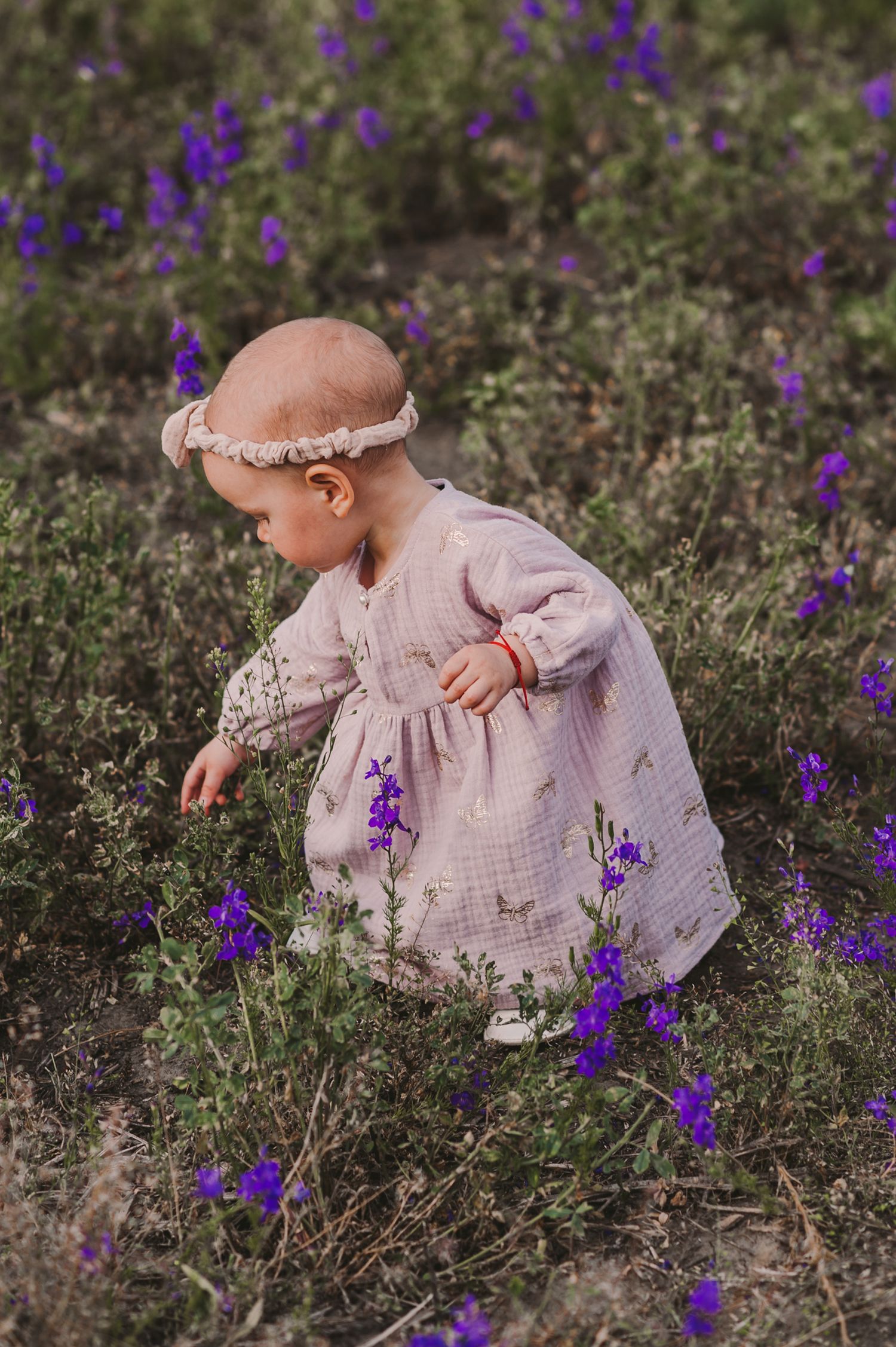A small child in a delicate dress explores a field of purple wildflowers on a sunny day.