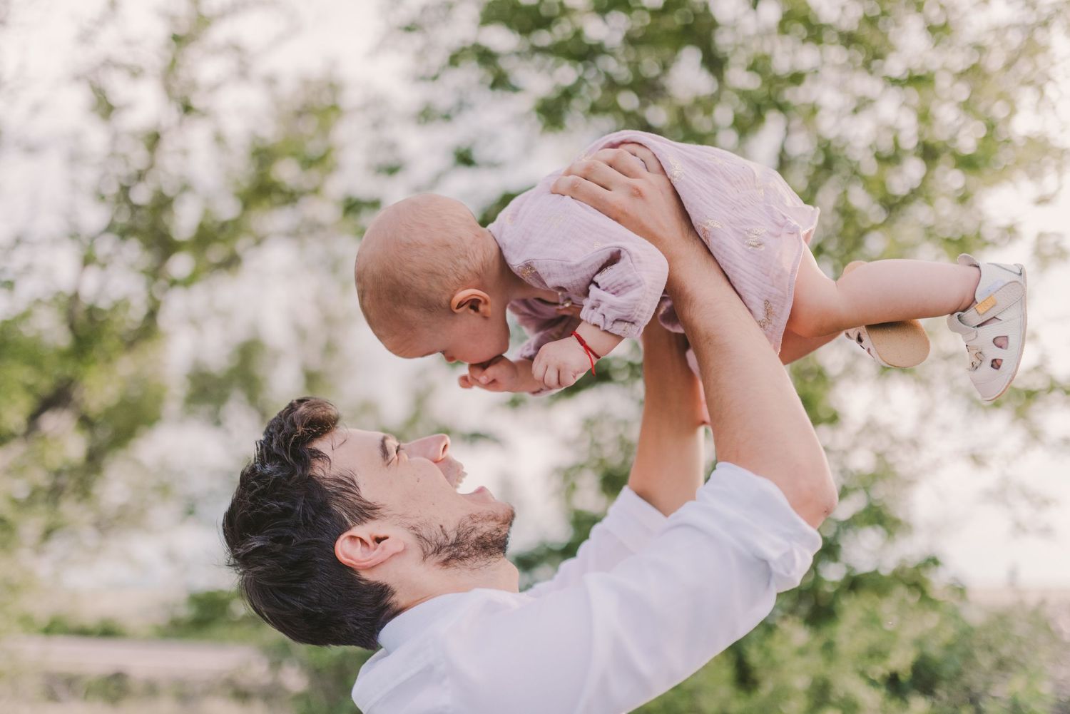 Parent playfully lifting baby in the air against a backdrop of green fields and trees.