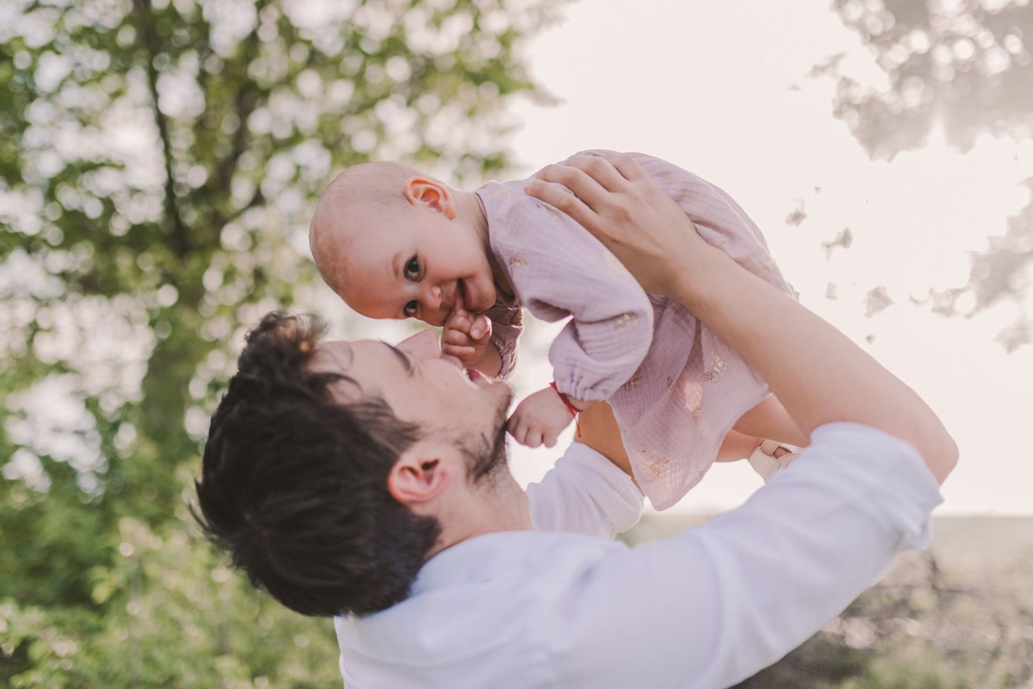 Parent lifting and playing with baby in white clothing against a natural outdoor setting.