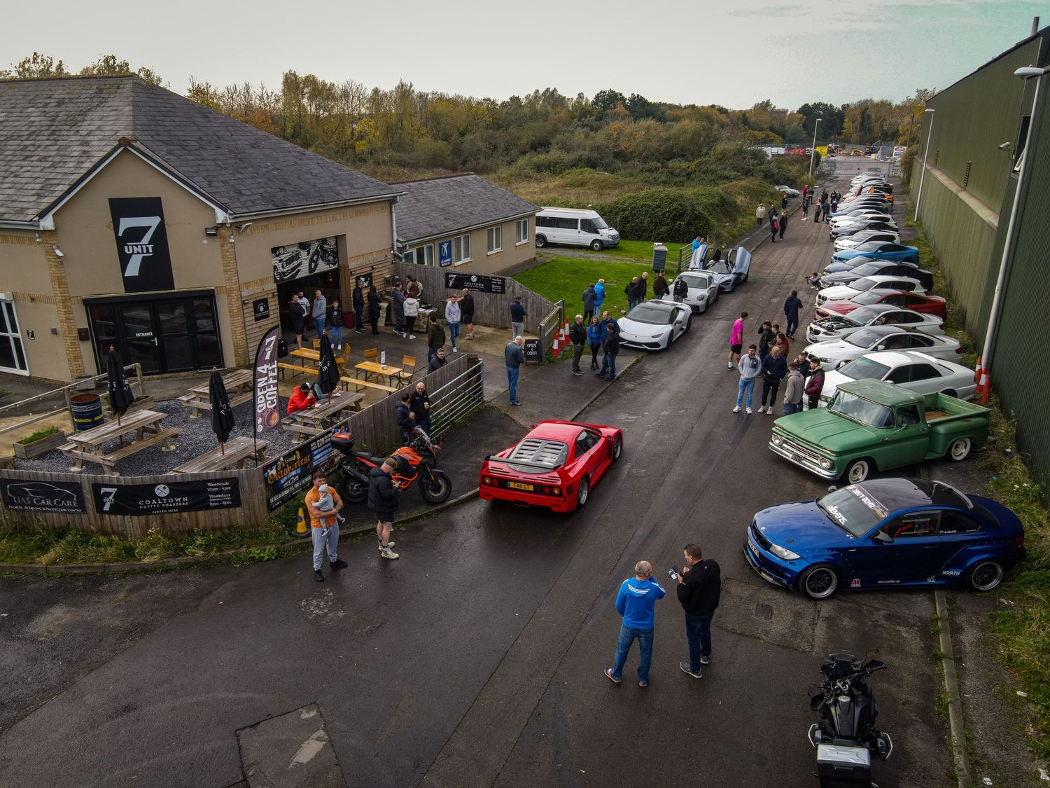 Aerial view of a car meet with vintage and modern vehicles lined up along a street.
