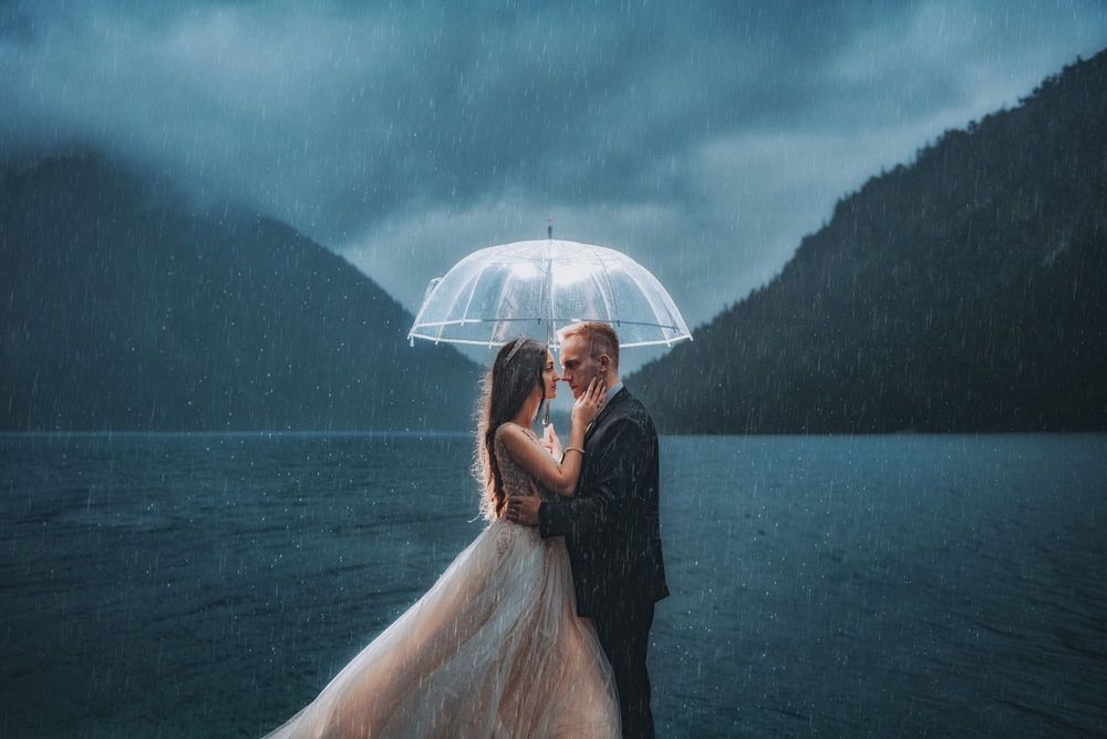 Wedding couple poses with clear umbrella against moody mountain lake backdrop.