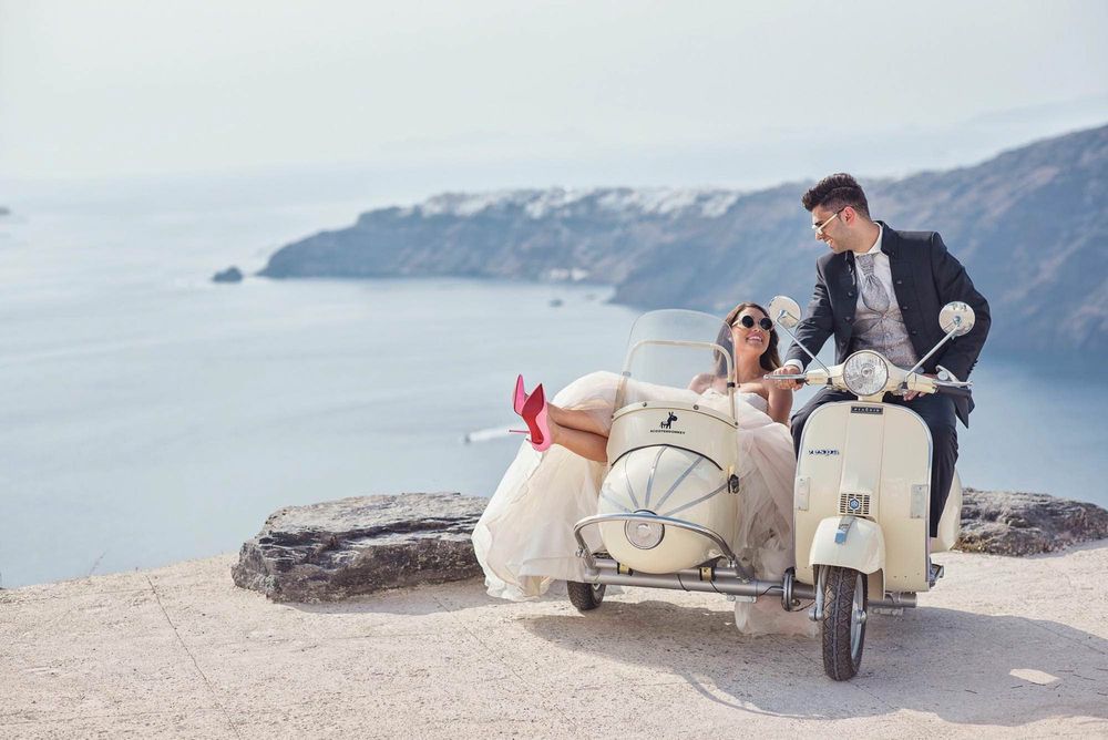 Wedding couple relaxes by vintage Vespa scooter on coastal Mediterranean road.