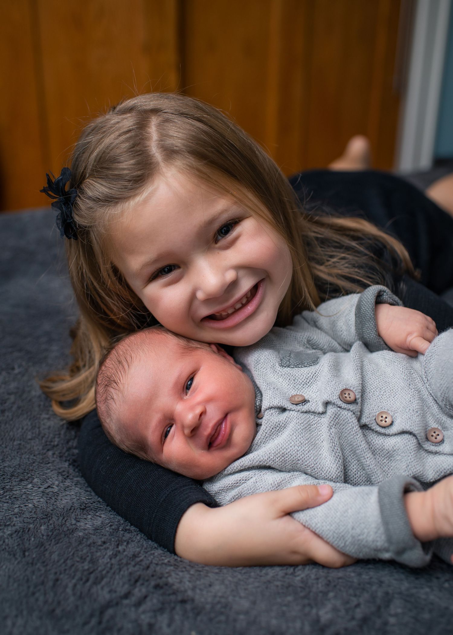 Two siblings share a tender moment as older sister cuddles with her newborn brother on gray couch.