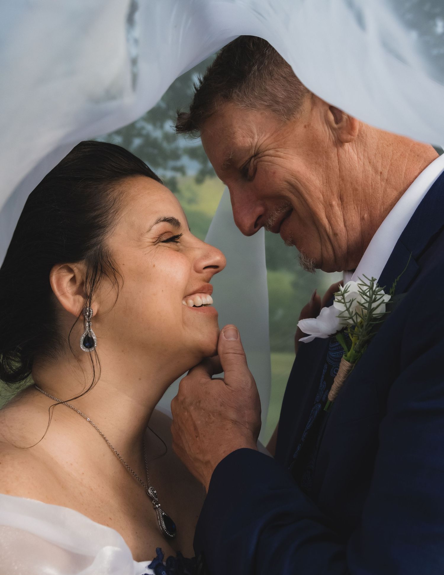 Bride smiling joyfully as groom gently holds her chin under a flowing white wedding veil.