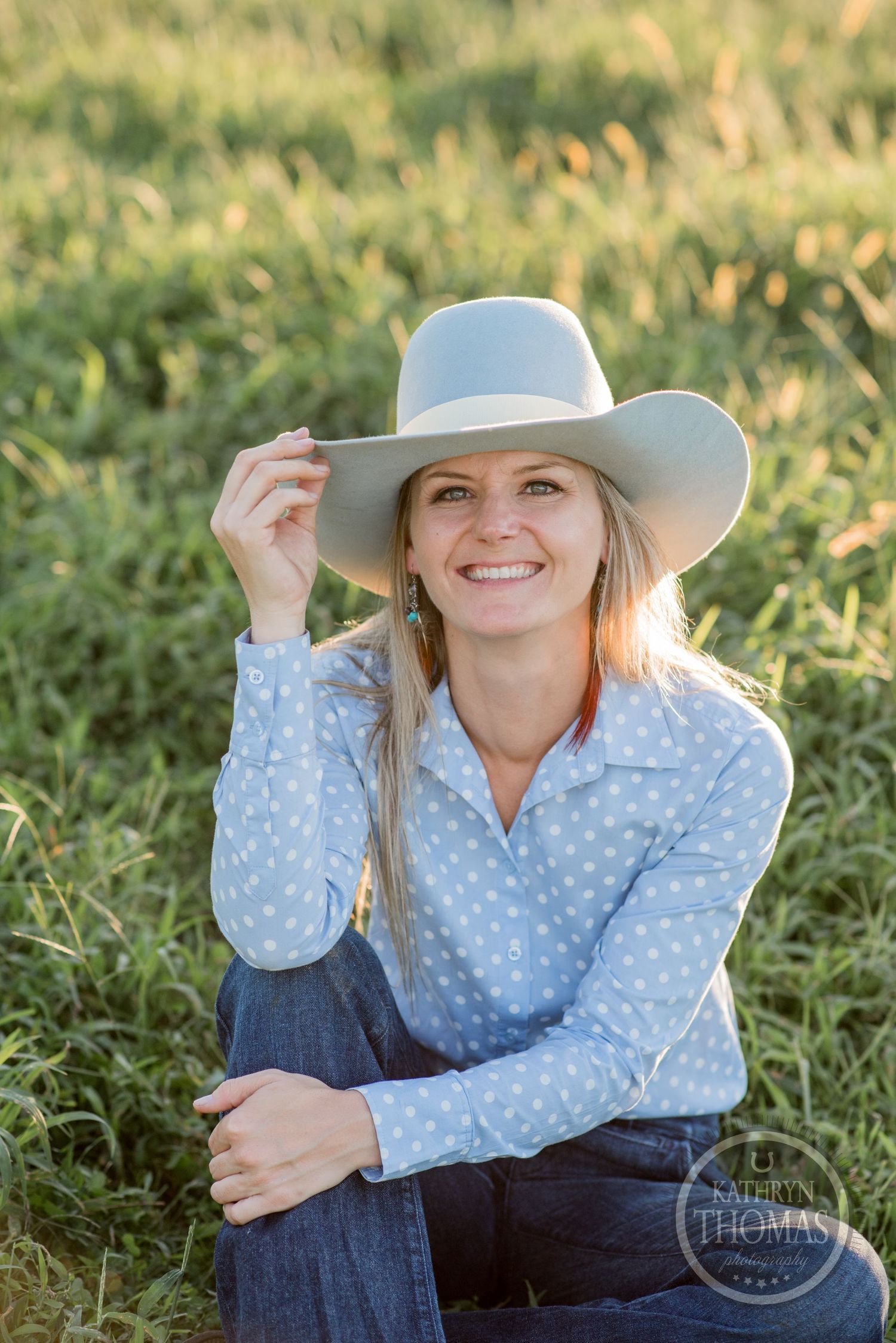 Someone in a polka dot shirt and cowboy hat sits in a grassy field during golden hour.