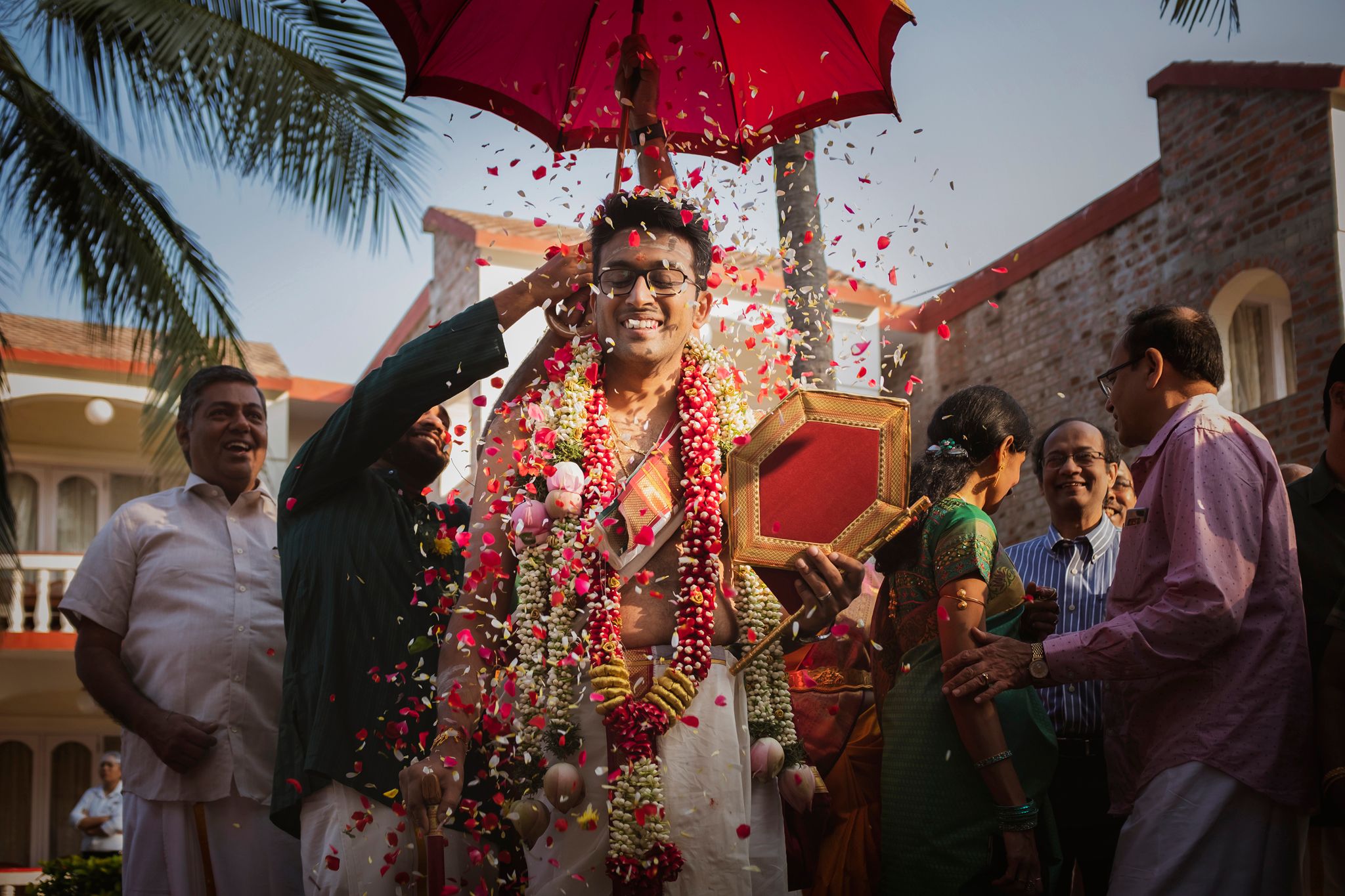 An Iyer wedding with flower petals being showered on the groom under a red umbrella during mappillai azhaippu or welcoming..