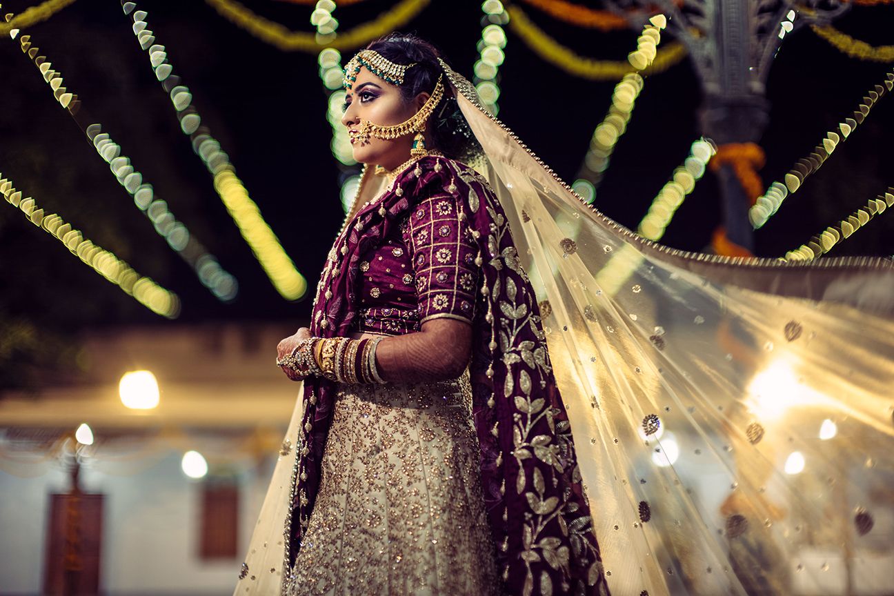 A bride in ornate traditional Indian attire with decorative lighting in the background.
