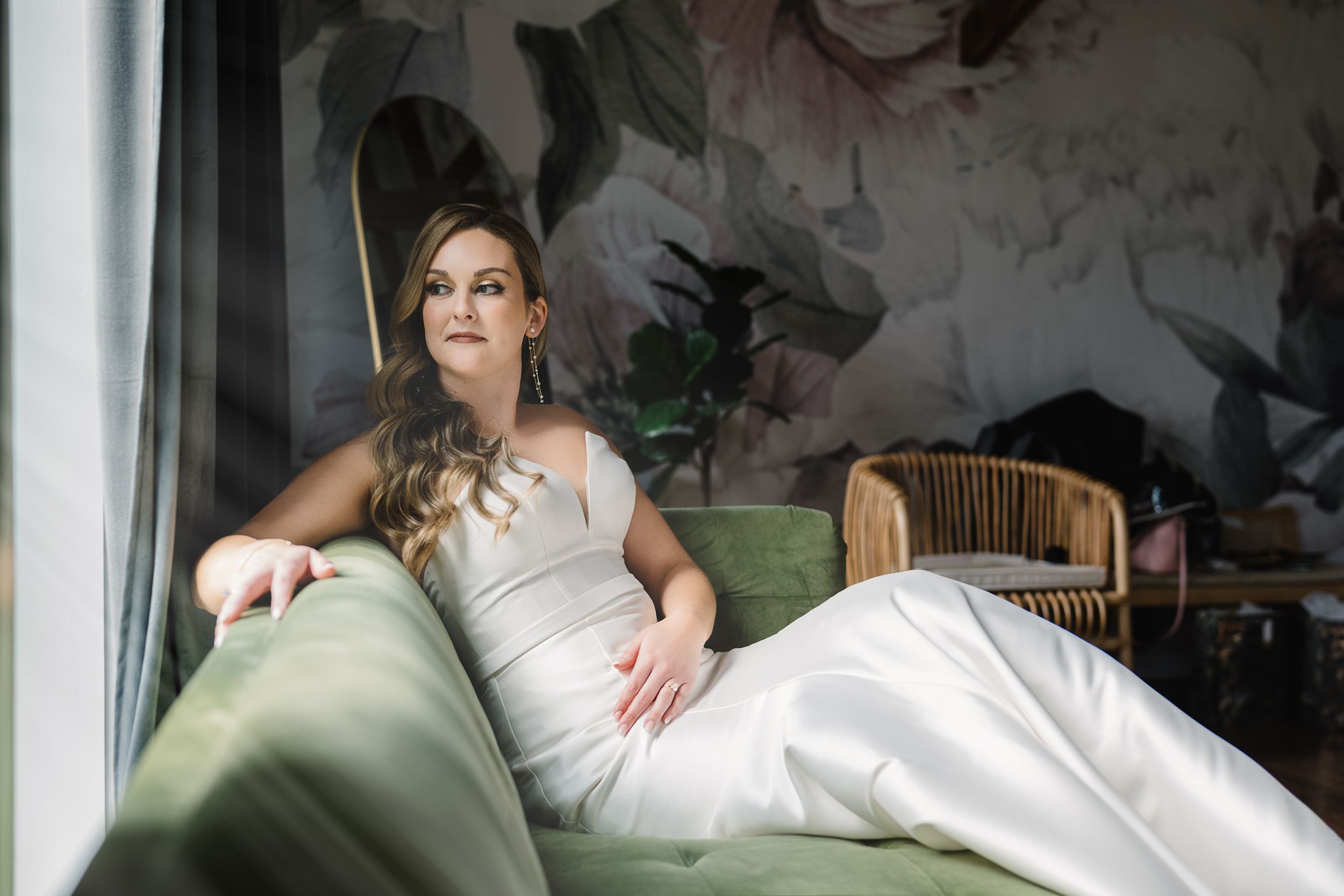 American bride in white dress reclining on green sofa near floral wallpaper.
