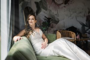 American bride in white dress reclining on green sofa near floral wallpaper, lit by soft window light in Austin.