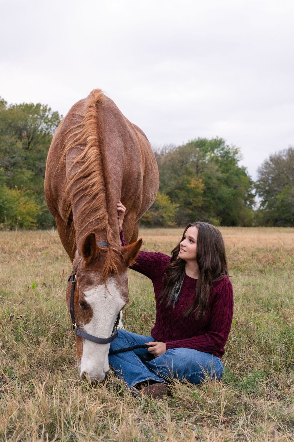 Horse Photoshoot in DFW, Texas. Girl sits cross legged and pets her horse while he eats grass in a large field on a cloudy day.