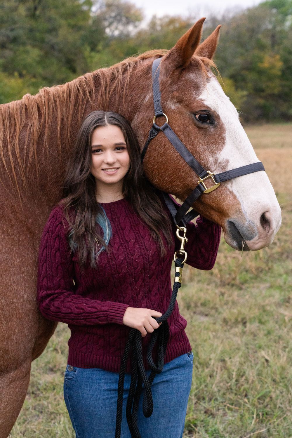 Horse photoshoot in DFW, Texas. Girl in a field stands in front her horse's neck and smiles while he looks to the side.