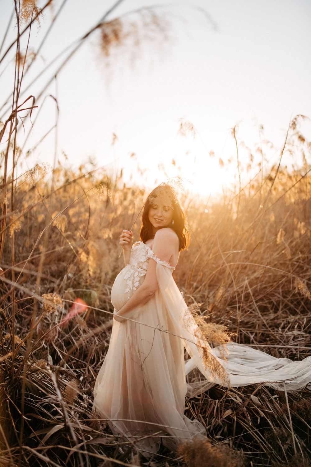 Ethereal maternity photo of an expectant mother in a flowing dress at golden hour among tall grasses.