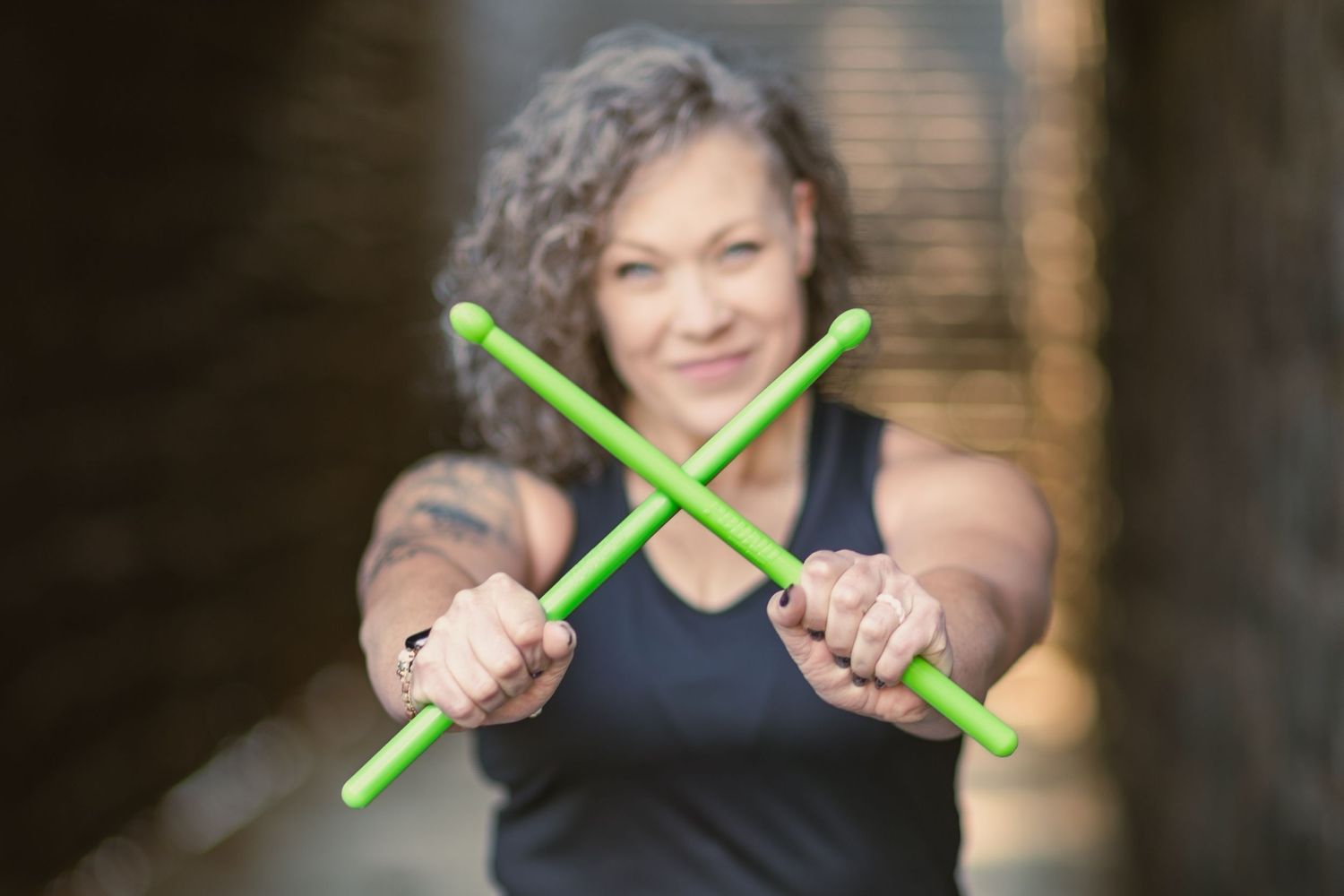 fitness woman holding drumsticks at camera in an x. she is in the background looking and smiling at camera. Oregon City