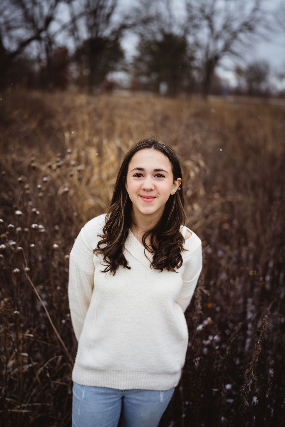 Portrait of a person in a cream sweater smiling outdoors against dried winter foliage.