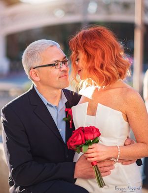 Romantic couple in wedding attire holding red roses against soft sunset backdrop.