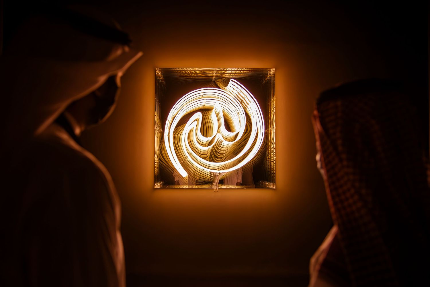 Illuminated Arabic calligraphic art piece glows orange between two viewing figures in traditional dress.