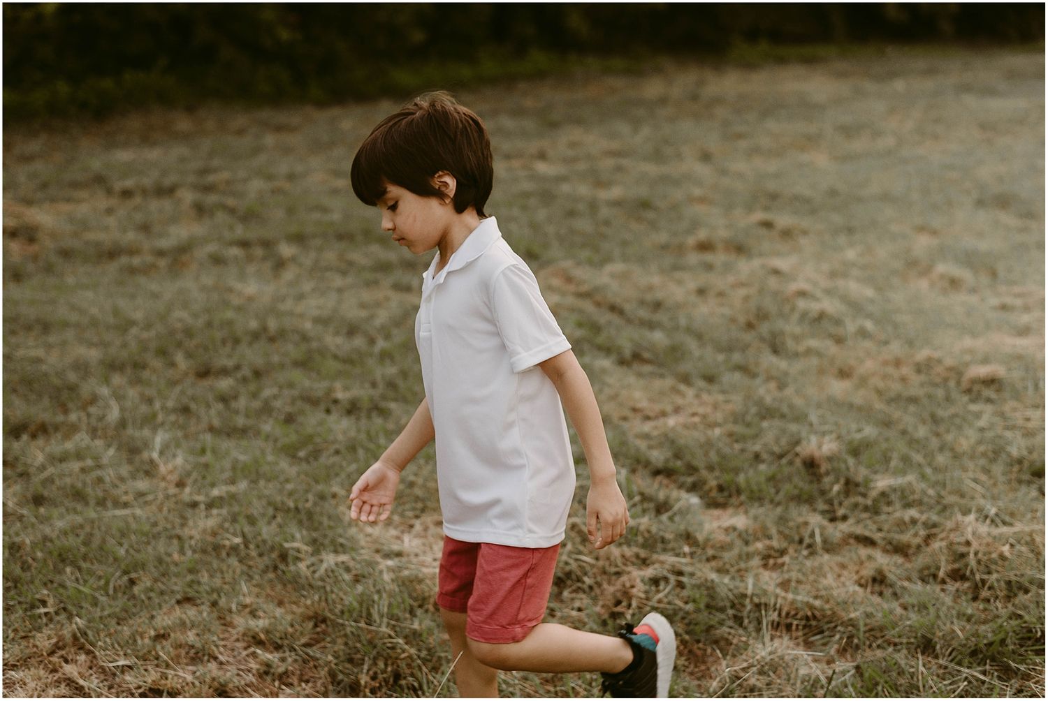 Child in white shirt and coral shorts runs through grassy field at sunset.