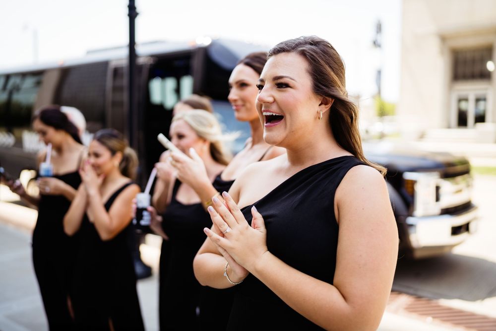 Bridesmaids in black dresses laugh and celebrate outdoors during a wedding celebration.