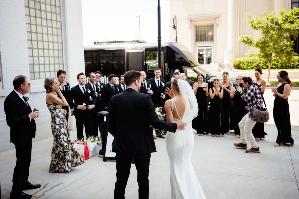 Wedding guests gather outside for a ceremony while a couple stands in front of them in formal attire.