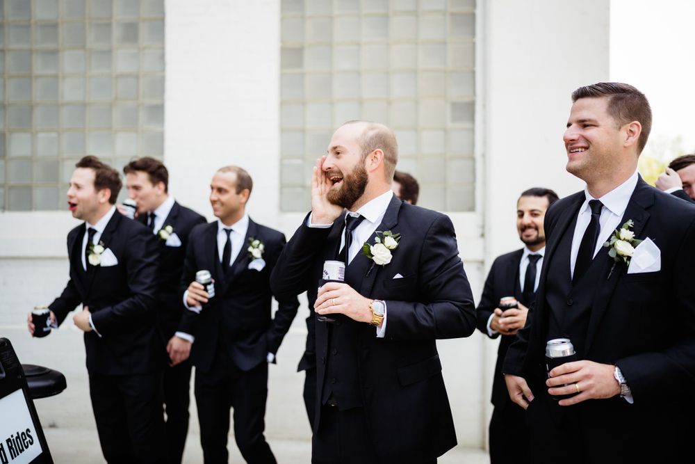 Groomsmen in black suits share laughs while holding drinks during a wedding celebration.