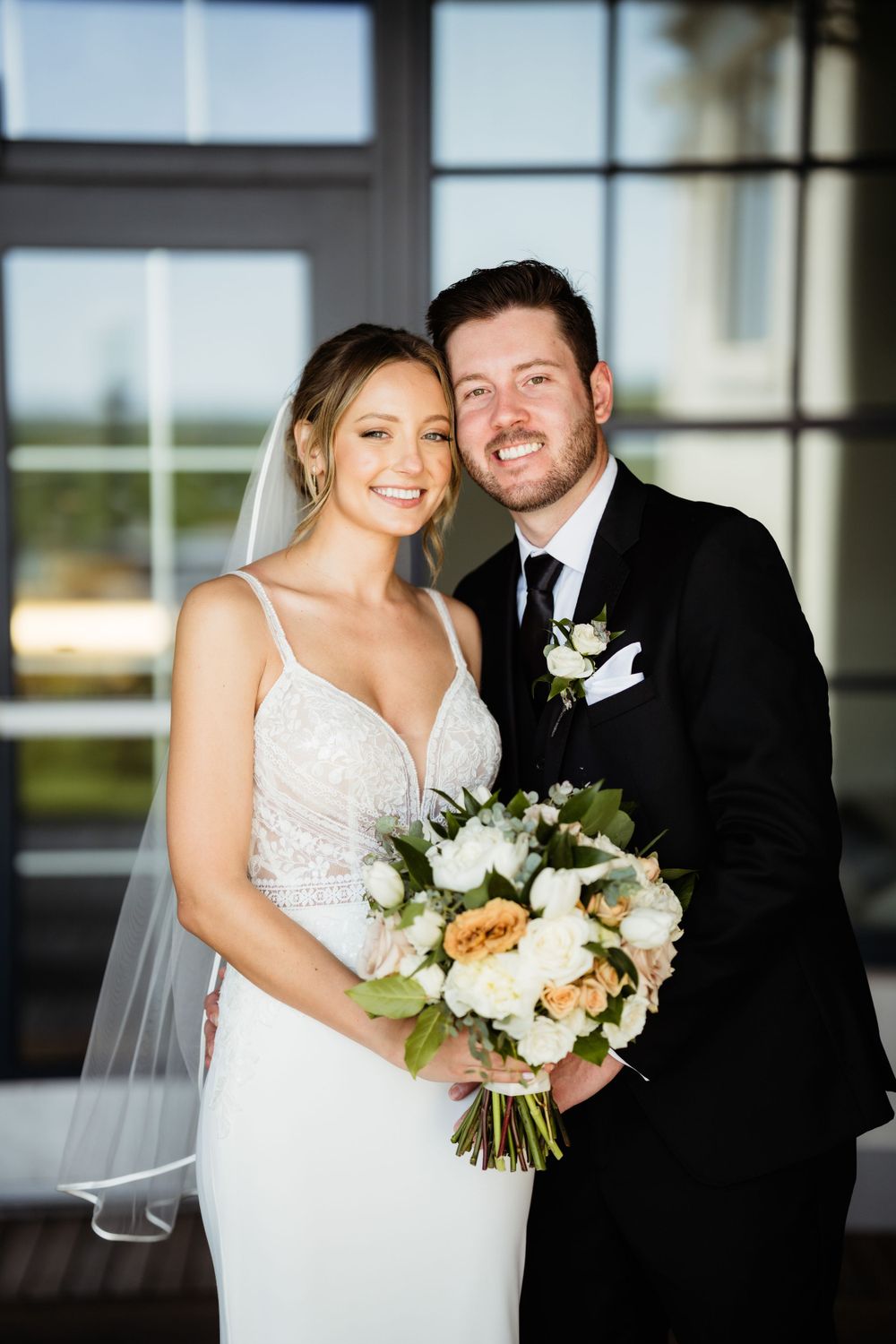 A wedding couple poses together indoors with a floral bouquet featuring white and peach blooms.