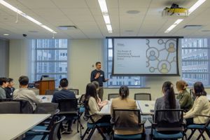 A presenter speaks to an audience in a modern classroom with a projection screen showing circular diagrams.