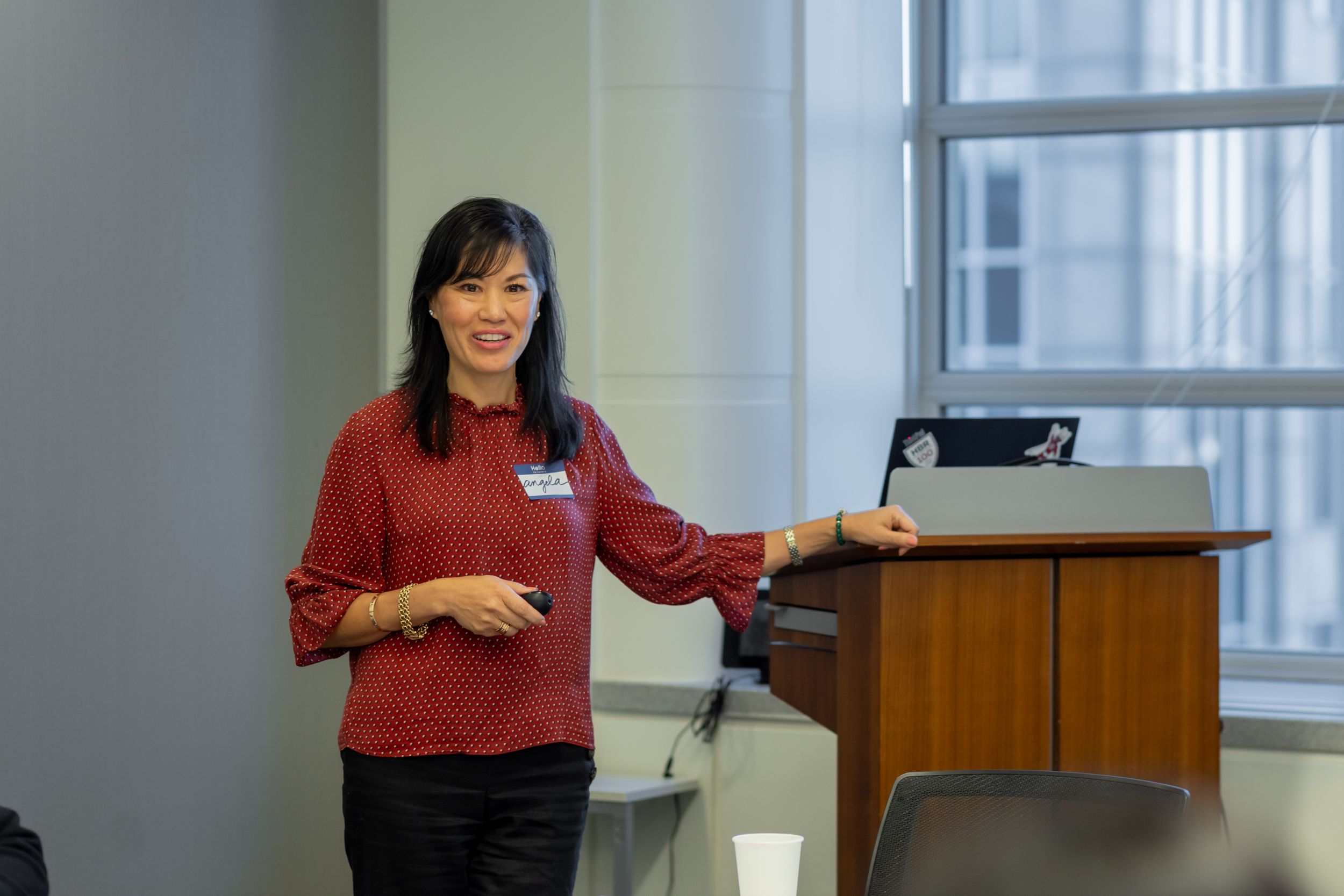 A speaker in a red sweater gives a presentation while standing at a podium near windows in an office setting.