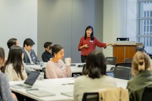 A classroom setting shows attendees taking notes while a presenter speaks near a projected presentation.