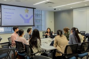 Meeting attendees seated at round tables face a presentation screen in a modern conference room.