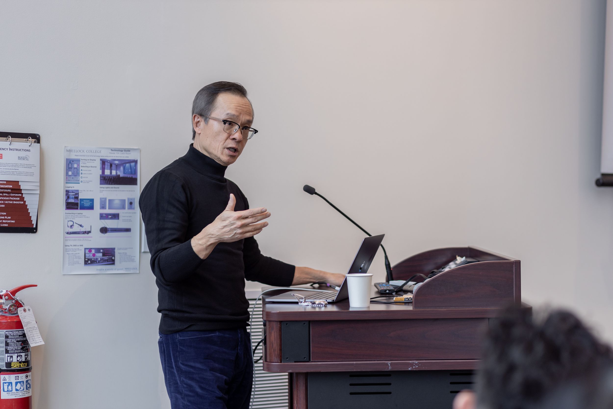 A presenter speaks at a podium during a professional event in a conference room setting.
