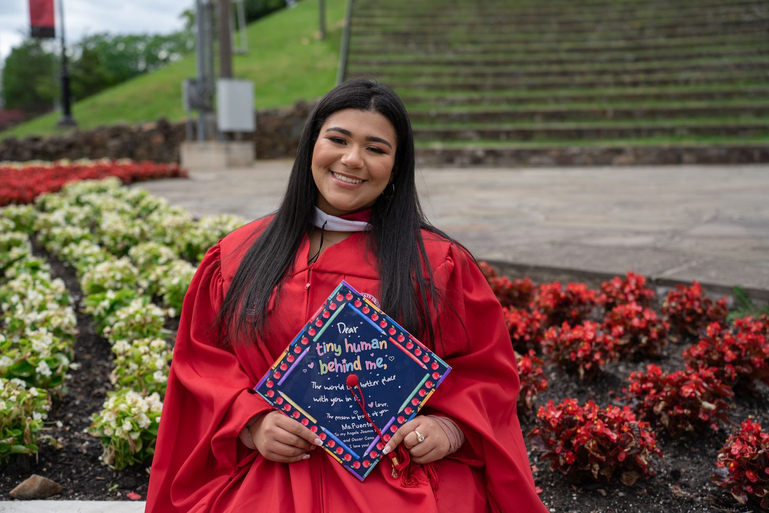Graduate displaying decorated cap and posing near red flowers on campus steps.