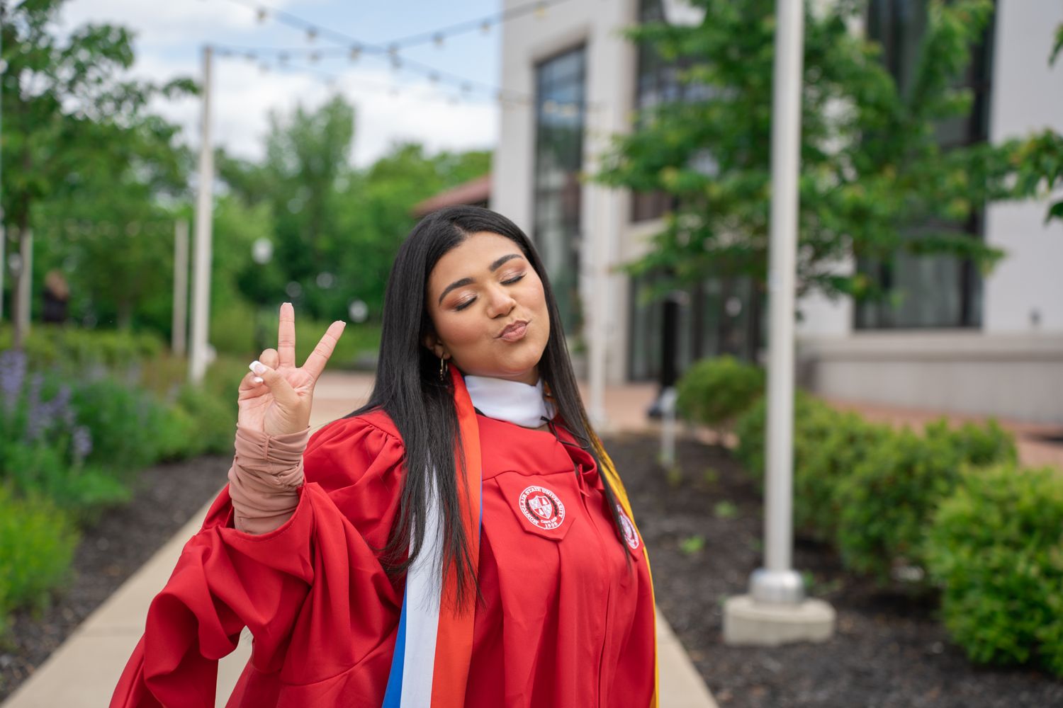 Graduate in bright red regalia celebrating on a walkway with decorative string lights.