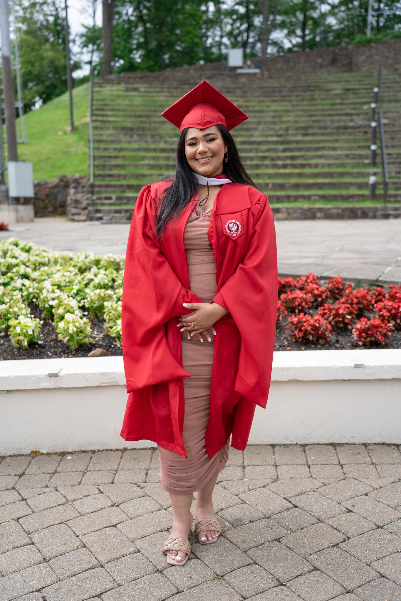 Graduate in red academic regalia posing on brick paver walkway with landscaped gardens.