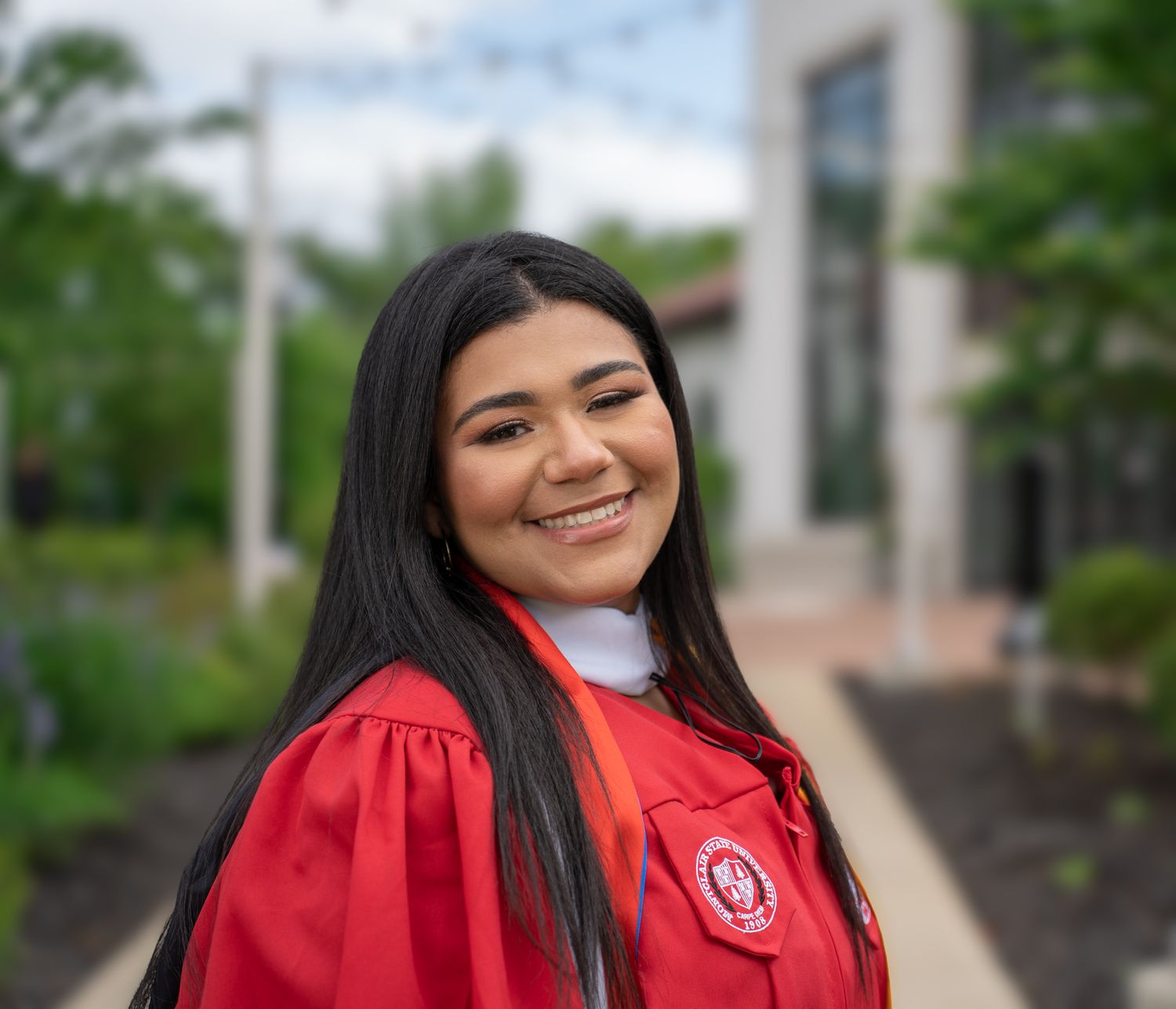 Graduate in red cap and gown posing outdoors on campus grounds.