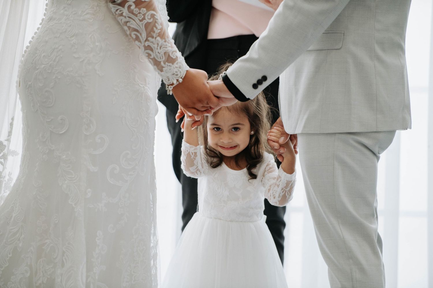 Young flower girl in white dress holds hands with the wedding couple during ceremony.