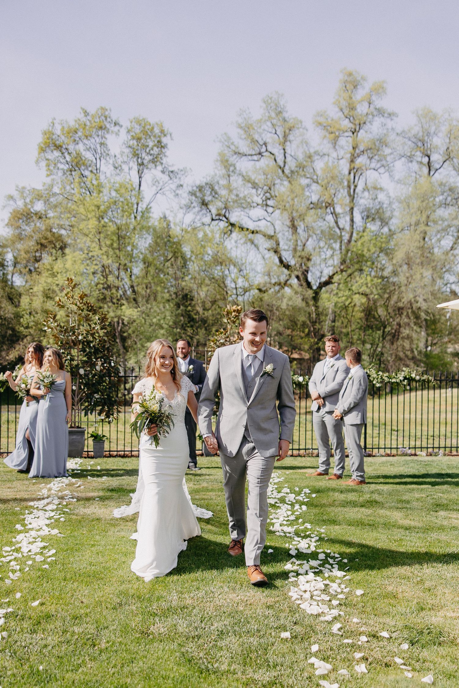 Wedding party proceeds down an outdoor aisle lined with white petals and surrounded by greenery.