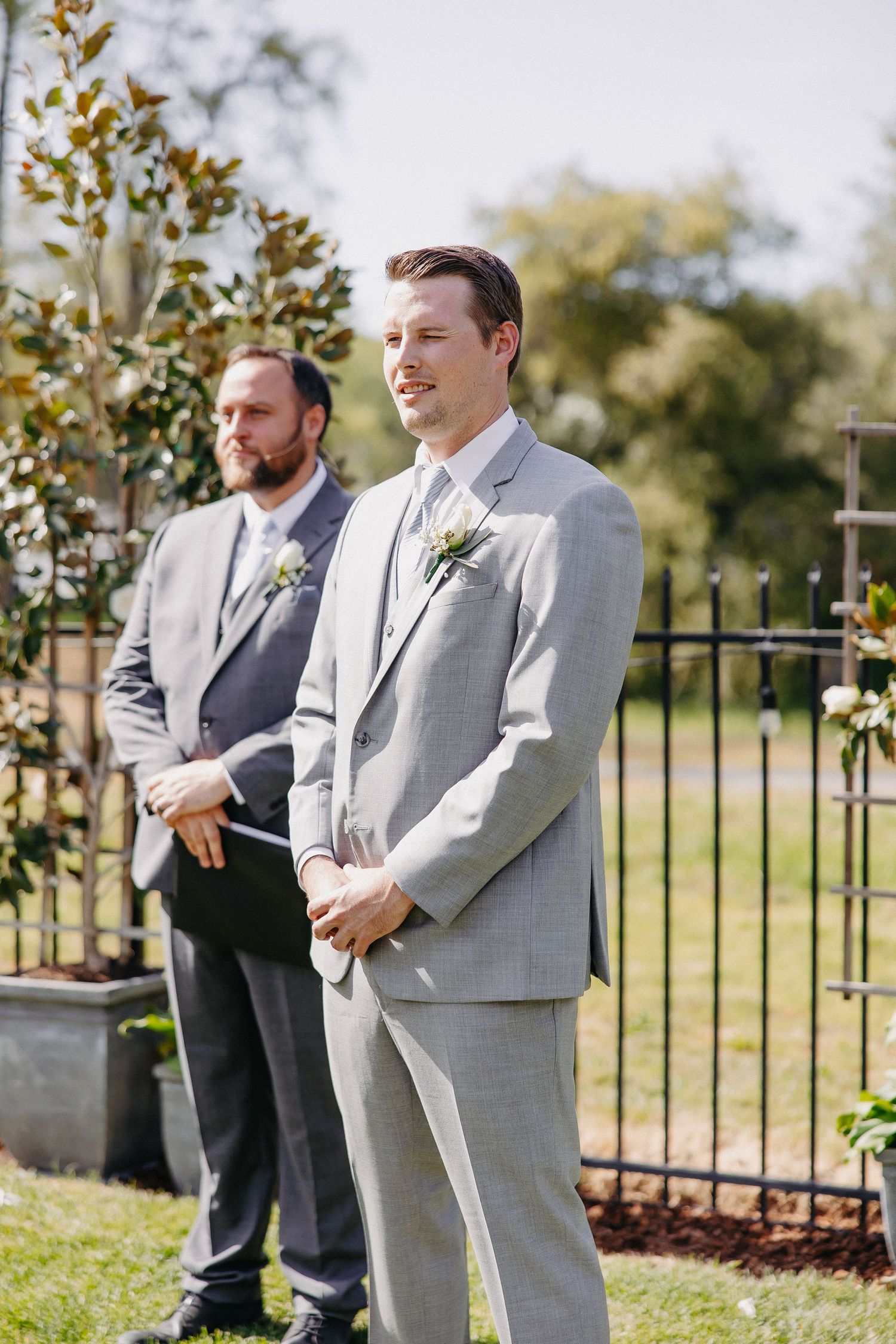 Two men in grey suits stand during an outdoor wedding ceremony.