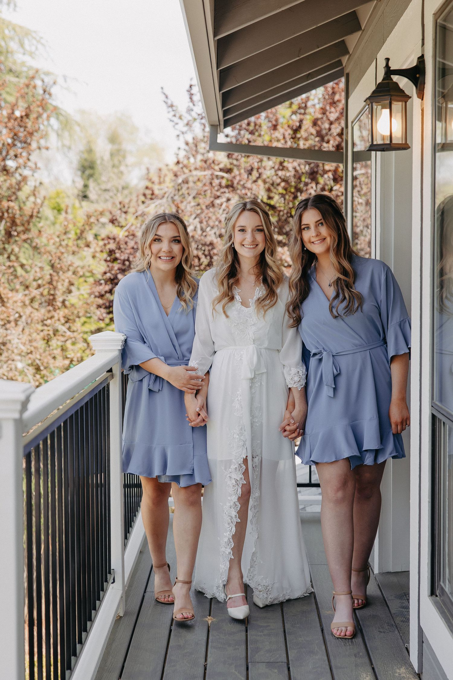 A bridal party in periwinkle blue dresses and white gown pose together on a deck with autumn foliage in background.
