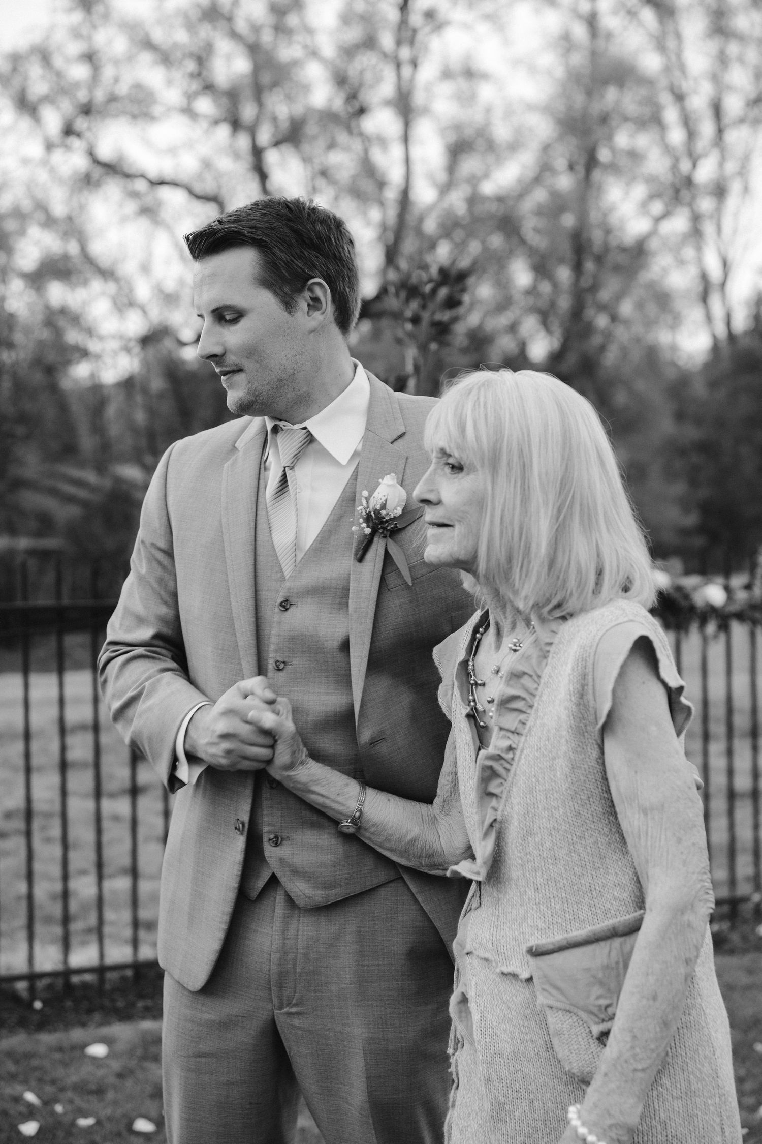 Black and white wedding photo of couple in formal attire standing by garden fence.