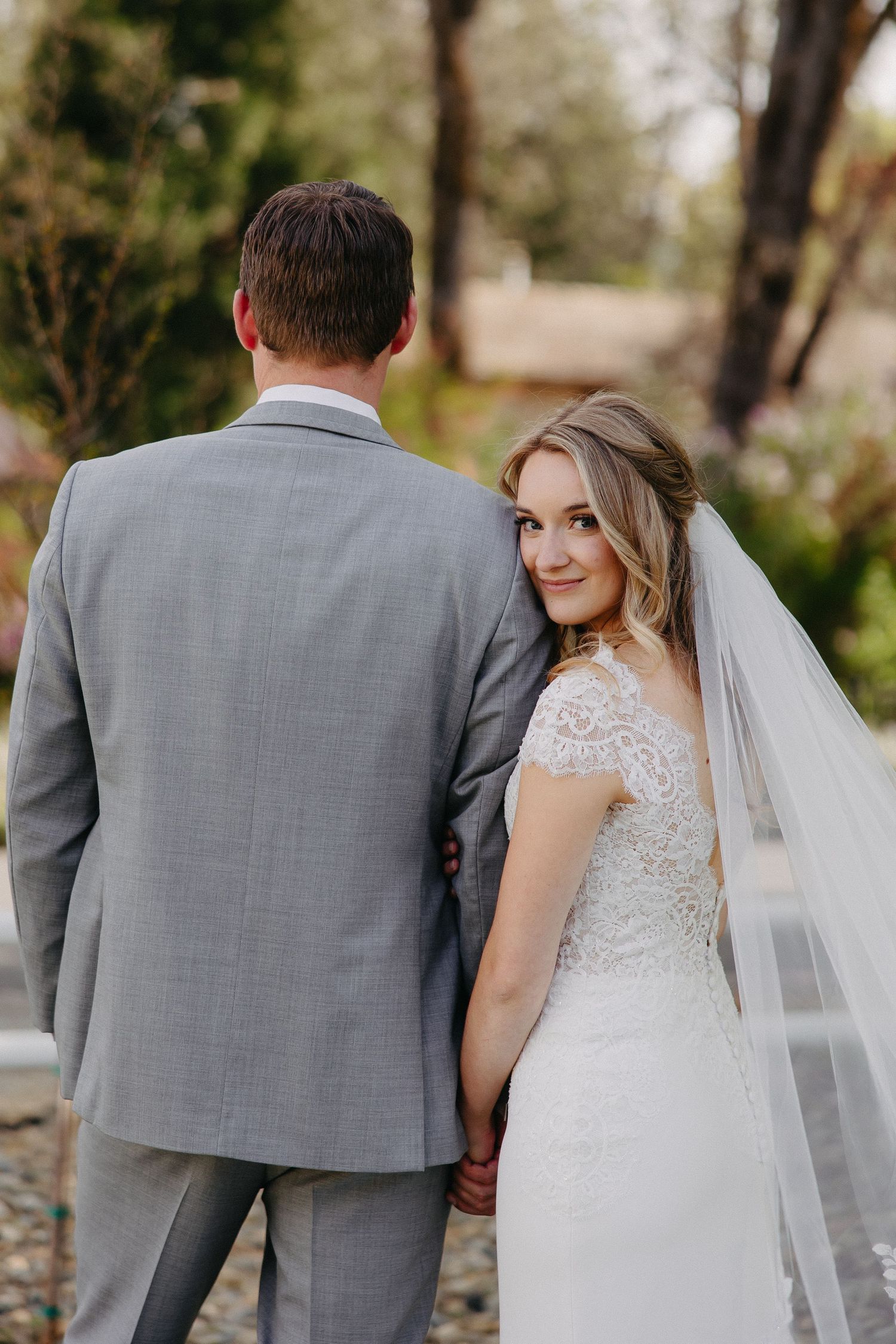 Wedding portrait capturing the bride's elegant lace dress and flowing veil from behind.