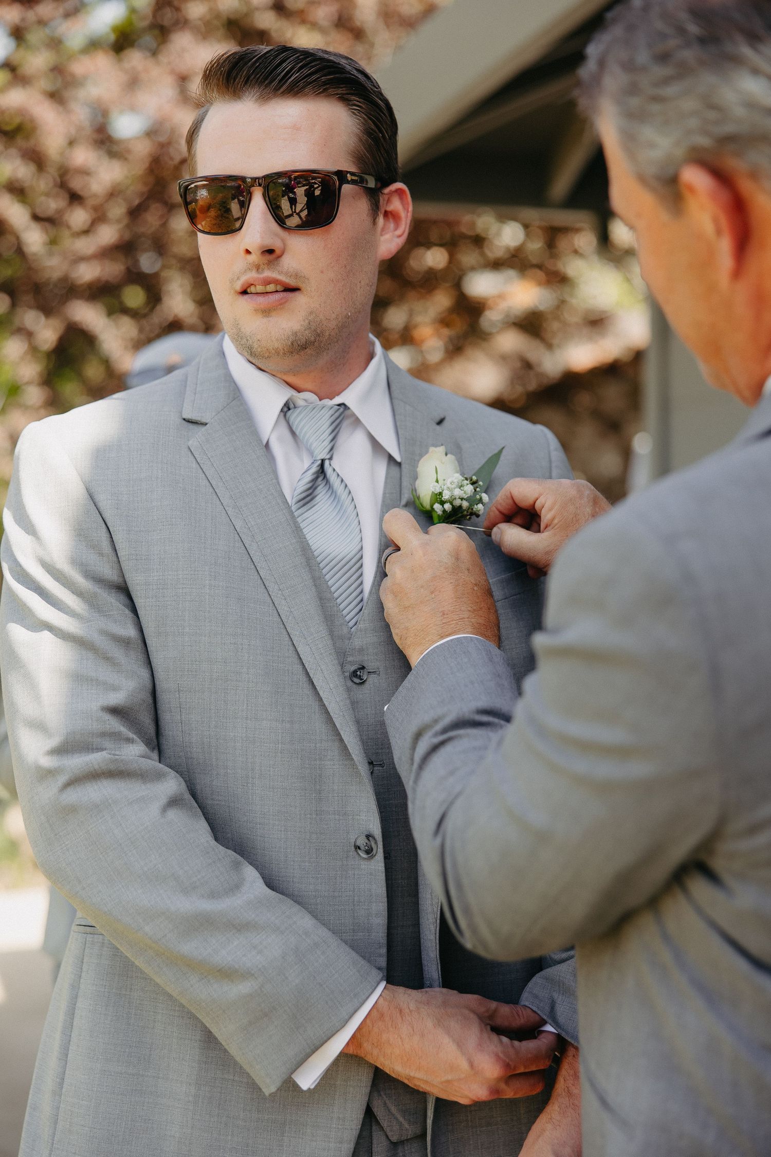 Groomsman in grey suit and sunglasses has boutonniere pinned to lapel outdoors.