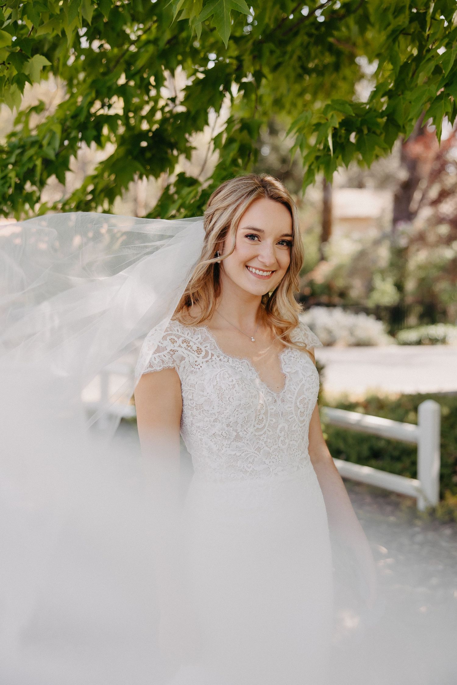 An ethereal bridal portrait with flowing veil captured beneath green maple leaves.