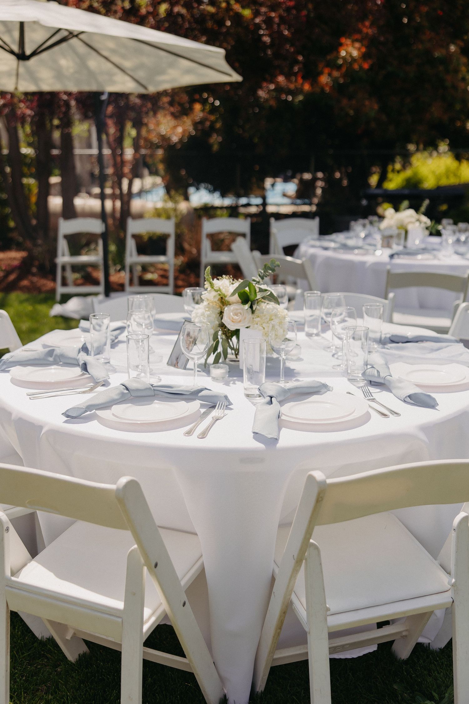 Outdoor wedding reception setup with white tables, chairs and umbrella in garden setting.