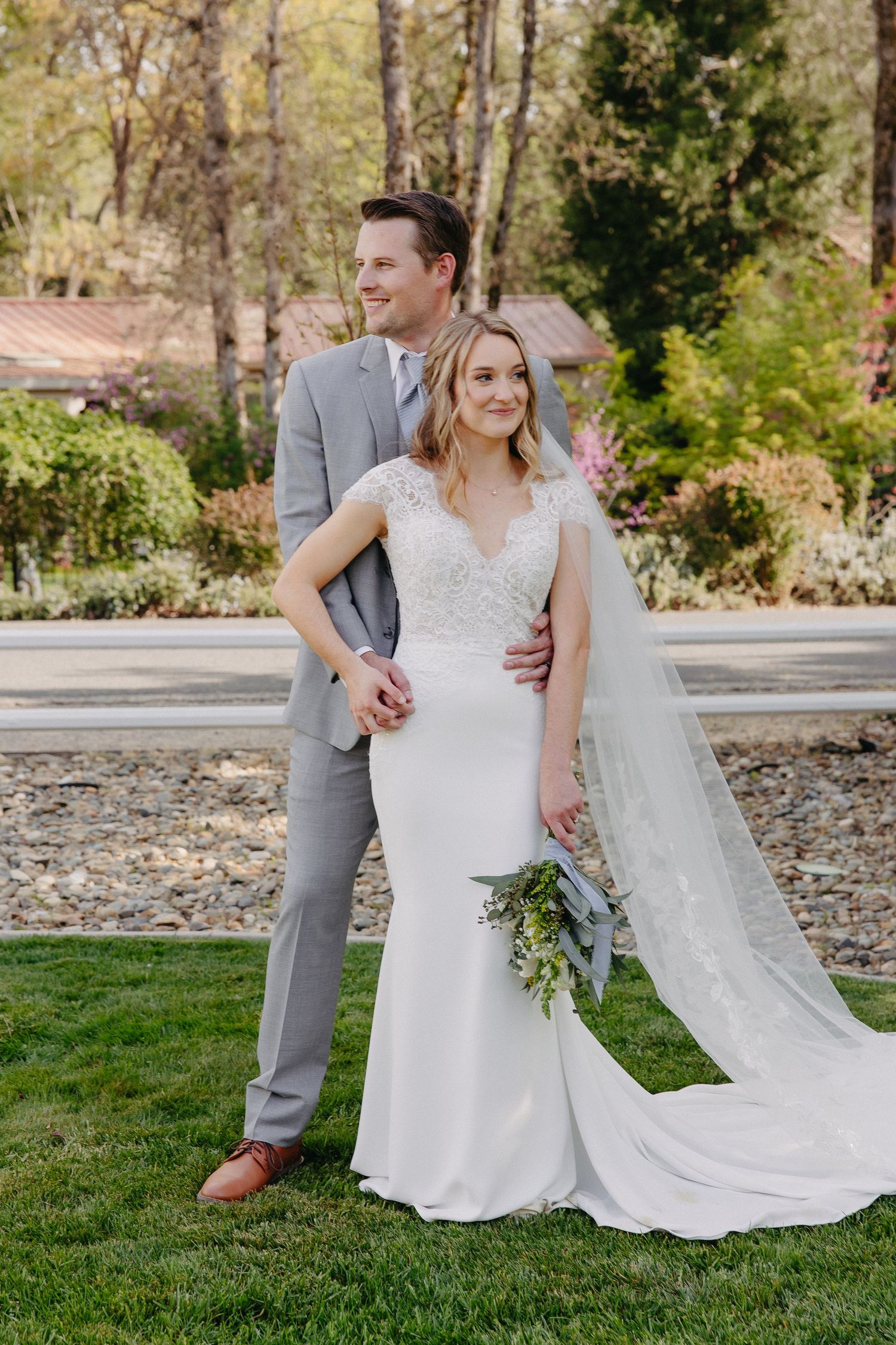 A couple poses elegantly in a garden setting, with the bride wearing a fitted lace gown and long veil.