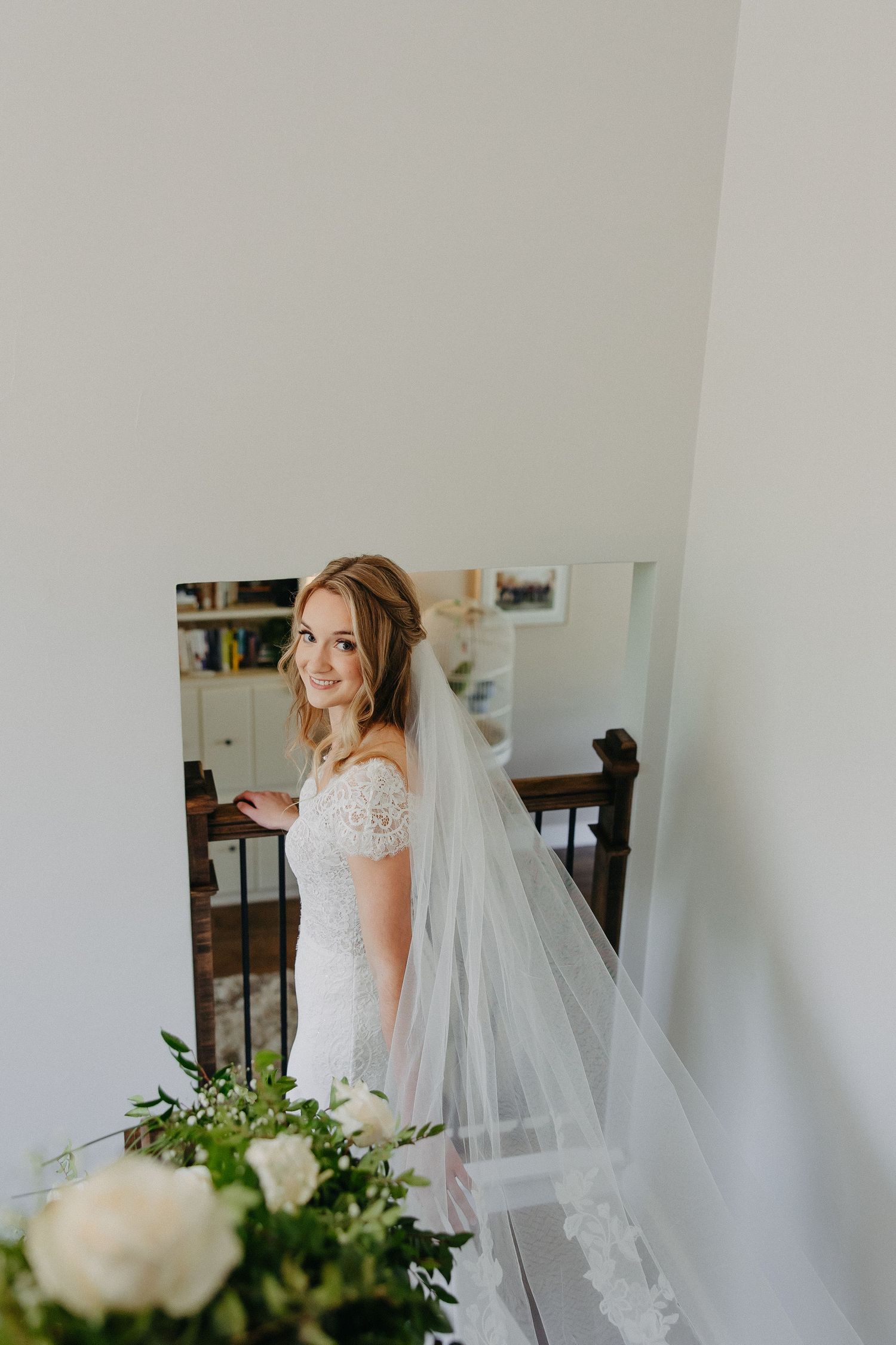 Radiant bride poses in a fitted lace wedding gown with flowing veil beside white floral arrangements.