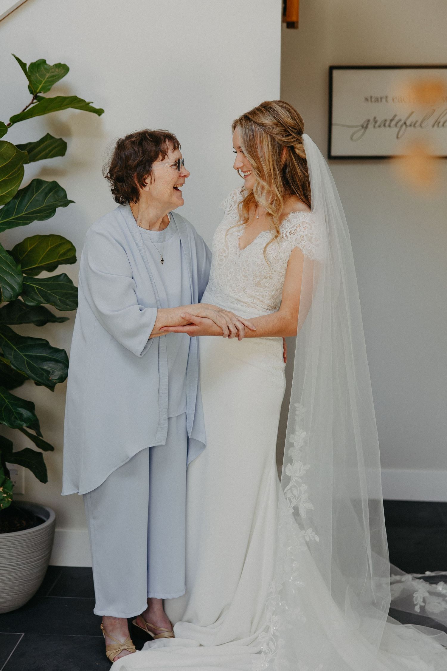 Tender moment between bride in lace gown and woman in light blue outfit beside indoor plant.