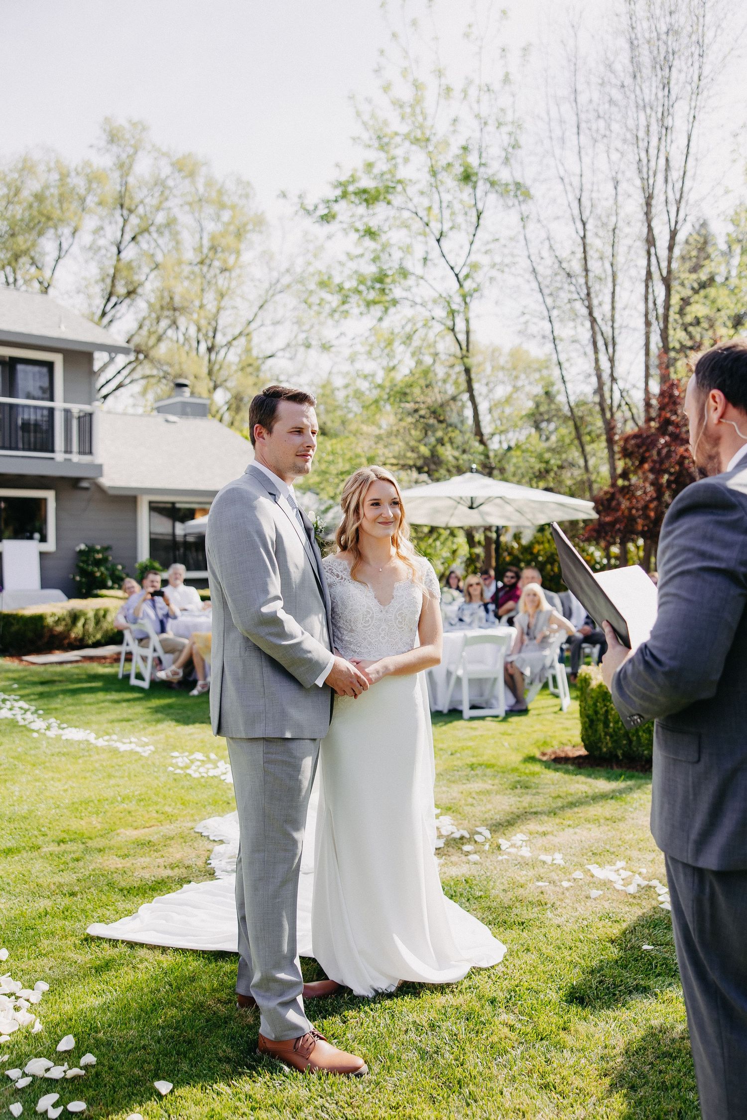 An outdoor wedding ceremony with guests seated on white chairs and flower petals lining the aisle.