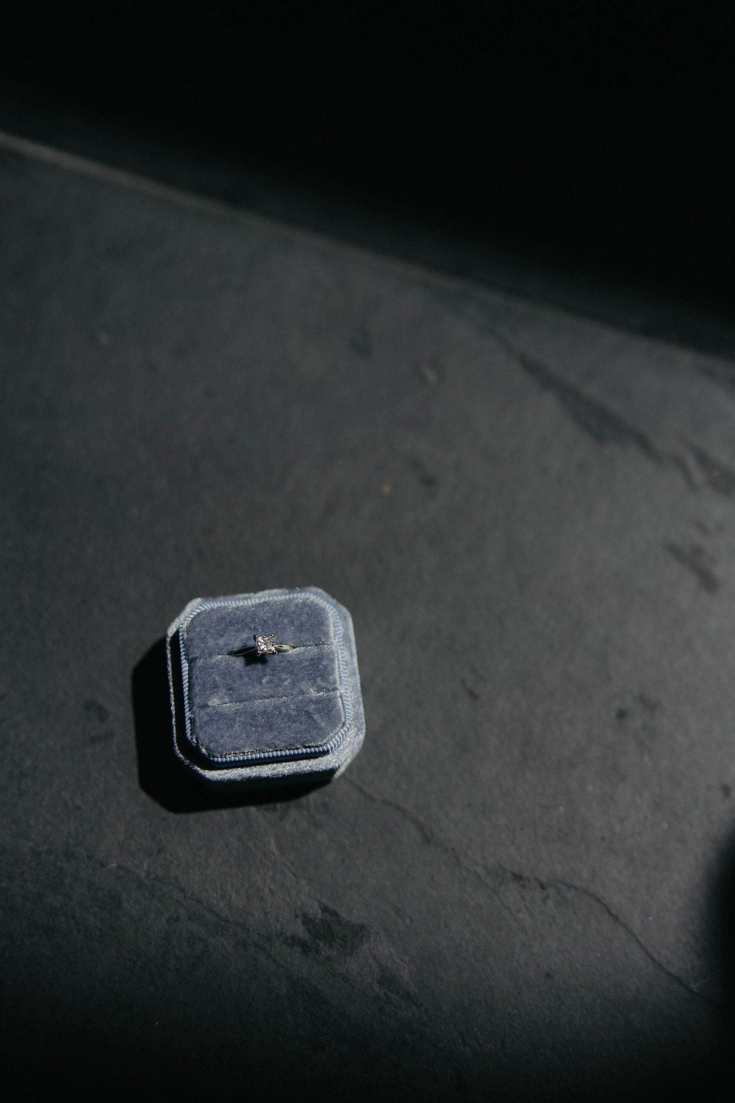 Single gray velvet ring box with engagement ring on dark moody surface.